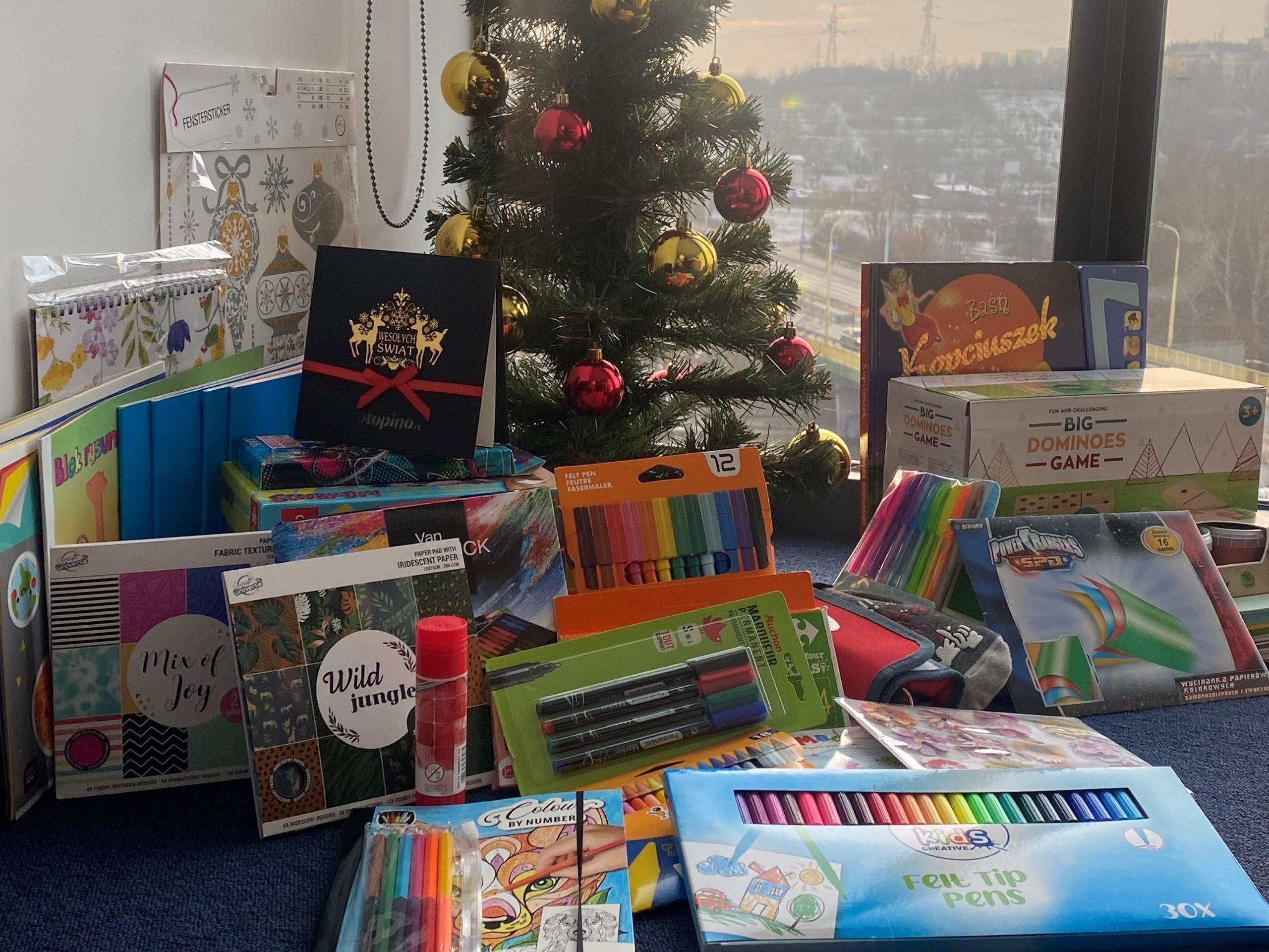 SU_2022_04_2 Upominki i materiały papiernicze pod choinką, z kartką firmy Rotopino.pl, w tle zachód słońca za oknem i widok na miasto