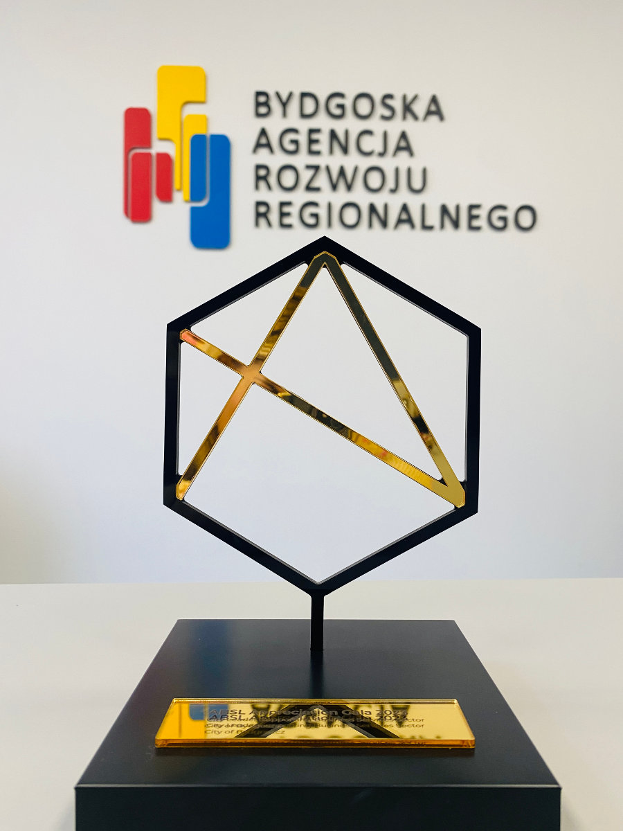 statuetka od ABSL statuetka, czarna kwadratowa podstawa, ze złotą blaszką z napisaną kategorią wyróżnienia, czarny sześcian na nóżce, w środku trzy złote linie łączące kąty sześcianu